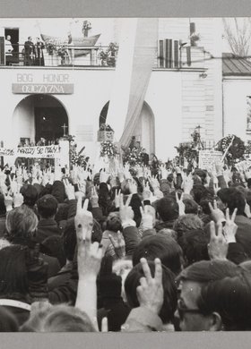 Pogrzeb bł. ks. Jerzego Popiełuszki - ludzie stoją przed kościołem p.w. św. Stanisława Kostki i trzymają prawe ręce w górze, a palce ułożone w literę "V"