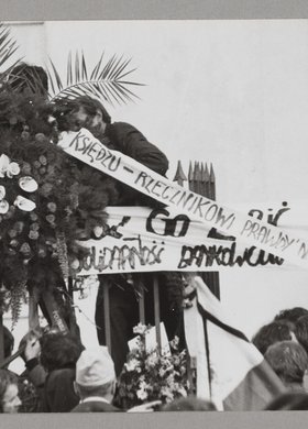 Pogrzeb bł. ks. Jerzego Popiełuszki - wieniec kwiatów leżący na katafalku z trumną ze zwłokami kapłana oraz liczne transparenty