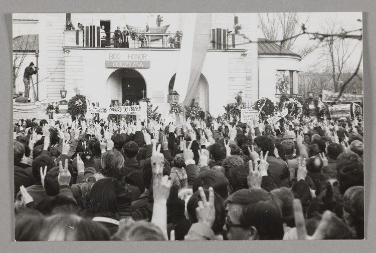 Pogrzeb bł. ks. Jerzego Popiełuszki - ludzie stoją przed kościołem p.w. św. Stanisława Kostki i trzymają prawe ręce w górze, a palce ułożone w literę "V"
