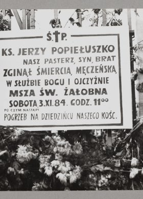 Pogrzeb bł. ks. Jerzego Popiełuszki - tablica przyczepiona do ogrodzenia kościoła p.w. Stanisława Kostki na Żoliborzu oraz wiązanki kwiatów