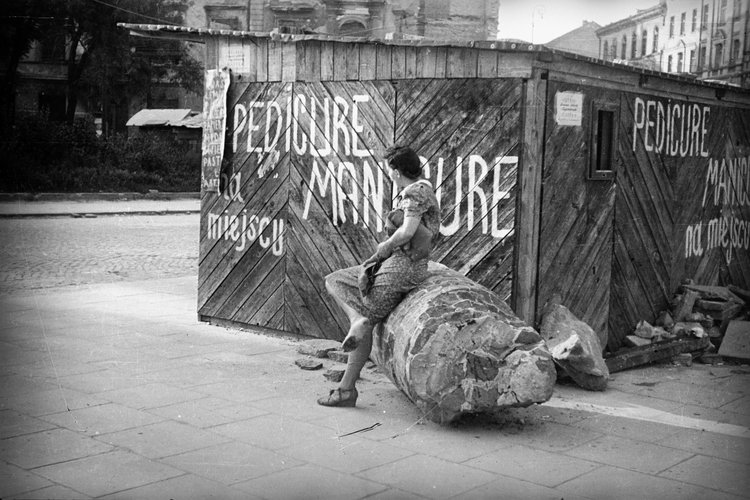 Maria Chrząszczowa siedząca na przewalonej kolumnie obok budki usługowej na Placu Trzech Krzyży