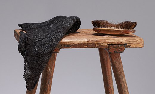 Fotografia przedstawiająca górną część drewnianego stołka, na którym leż ciemna tkanina oraz szczoteczka do włosów