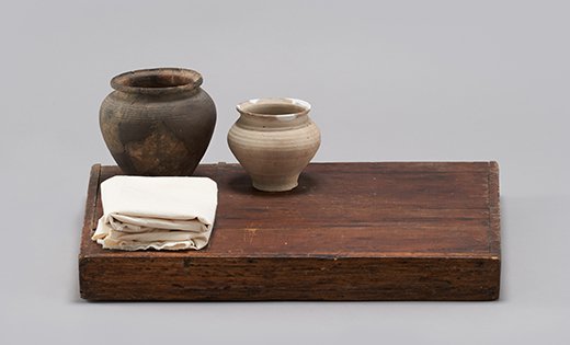 Fotografia przedstawiająca drewnianą stolnicę, na której po lewej stronie ustawione są w rzędzie dwa naczynia ceramiczne. Przed naczyniami leży złożona piała tkanina