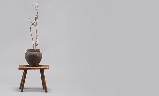 Fotografia przedstawiająca drewniany stołek ustawiony po lewej stronie. Na nim stoi gliniane naczynie, w którym znajdują się suche gałązki.